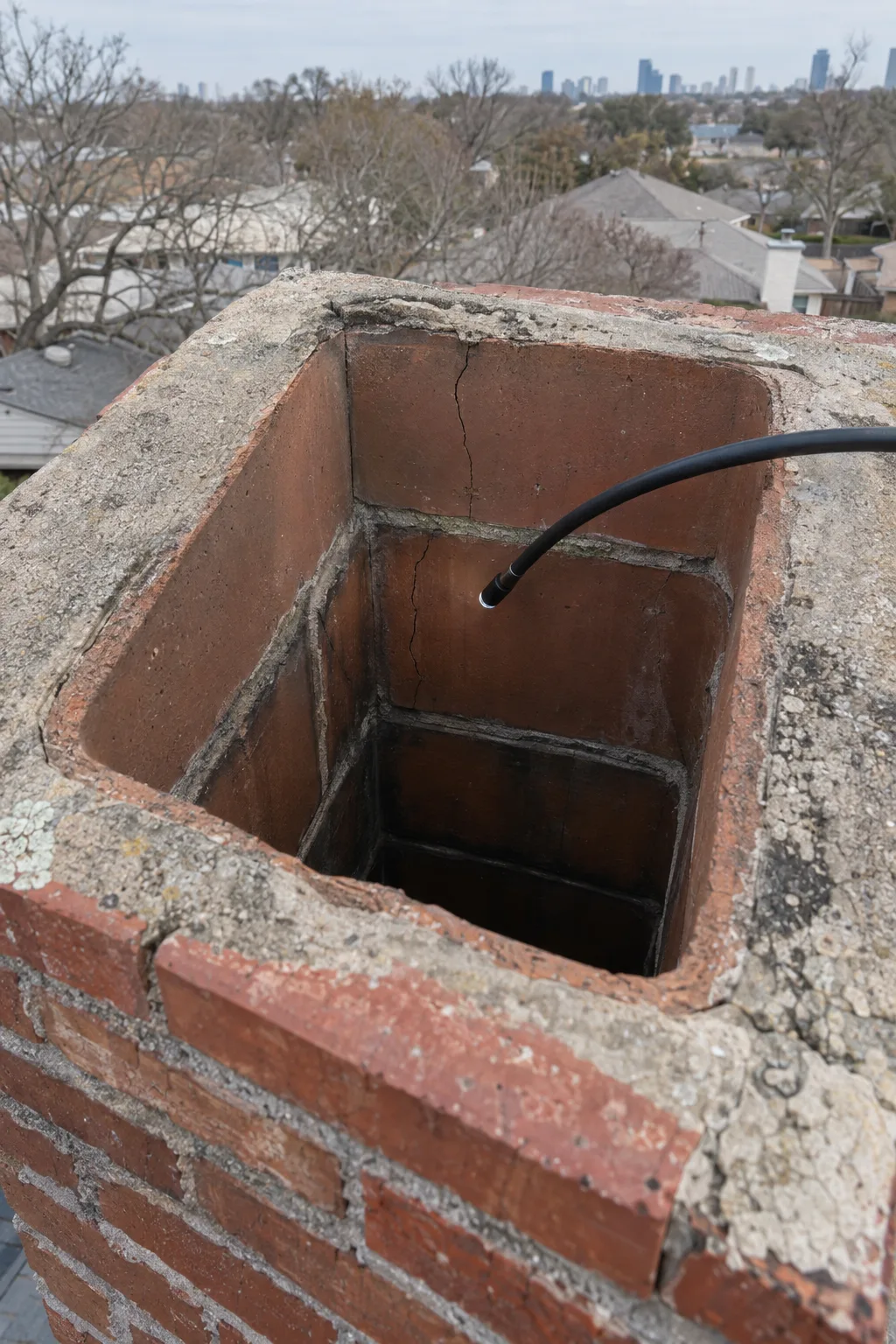 View from above into a brick chimney flue showing cracked clay tile liner sections and deteriorated mortar joints, with a camera inspection cable inserted into the opening and a sun-bleached Dallas suburban roofline in the background.