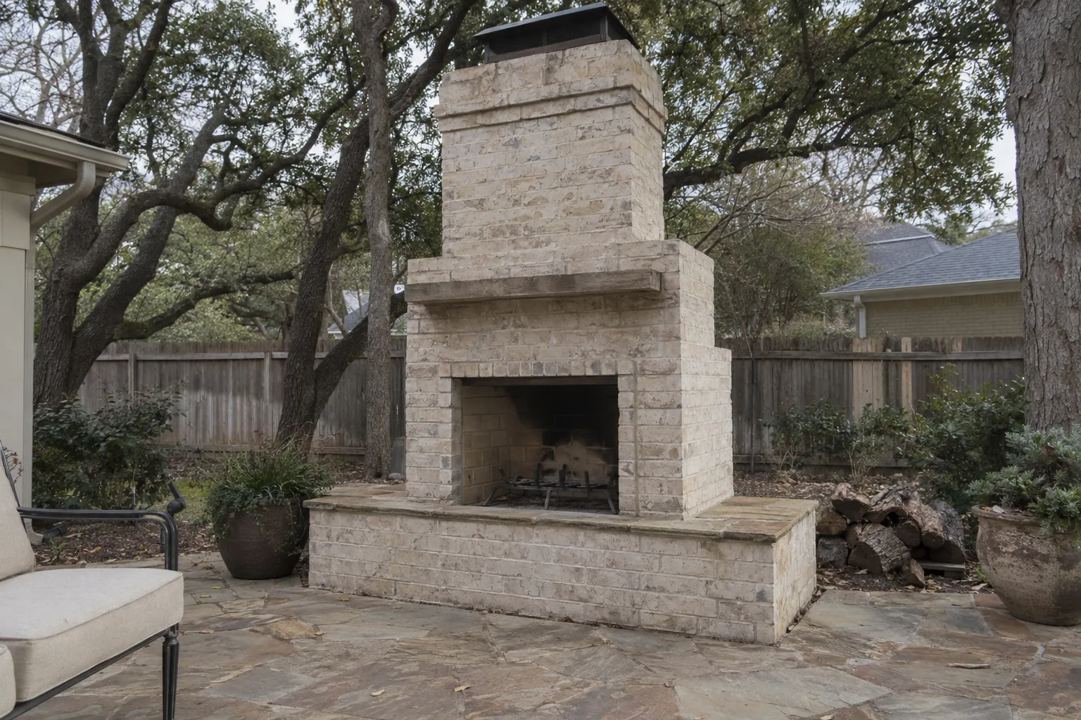 A sturdy masonry outdoor fireplace with a brick surround and open firebox sits on a flagstone patio in a Dallas backyard, surrounded by live oak trees in warm afternoon light