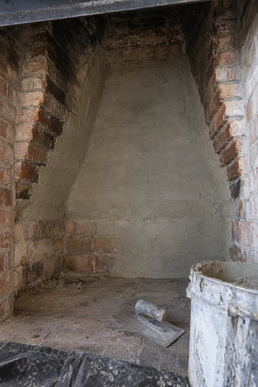 Interior of a brick fireplace smoke chamber with freshly applied smooth grey refractory parging mortar coating the corbeled walls, a trowel and mortar bucket resting on the hearth below